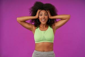 Posititve young curly dark skinned female in light green top posing over purple background with palms on her head, smiling widely to camera and demonstrating her white perfect teeth photo