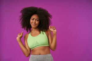 Studio photo of young dark skinned lady in sporty clothes standing over purple background, looking down and frowning confusedly with raised hands