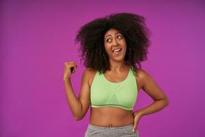 Joyful young sporty dark skinned female with curly hair isolated over purple background, looking aside with confused face and raising hand, wearing light green top photo