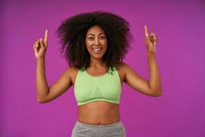 Portrait of young beautiful dark skinned female with curly hair showing her pleasant emotions, pointing upwards with index fingers and looking at camera happily, isolated over purple background photo