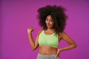 Portrait of curly dark skinned lady with casual hairstyle wearing light green top, smiling slightly to camera with raised fist, posing over purple background photo