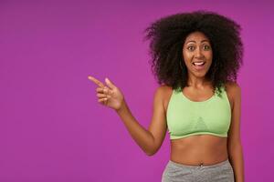 Happy young attractive dark skinned female with curls wearing light green top, pointing aside with forefinger and looking at camera with cheerful wide smile, standing over purple background photo