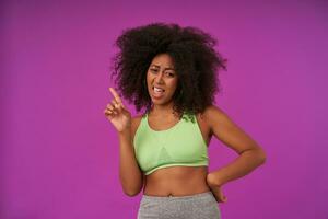 Portrait of young curly woman with dark skin in light green top standing over purple background, looking down wide mouth opened and raising forefinger up photo