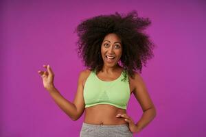 Beautiful female showing her pleasant emotions over purple background, wearing light green sporty top, smiling joyfully with wide mouth opened, keeping one palm on waist and raising other up photo