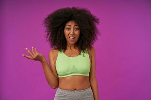 Puzzled young curly female with dark skin wearing casual hairstyle, raising palm perplexedly and looking to camera with raised eyebrows, isolated over purple background photo