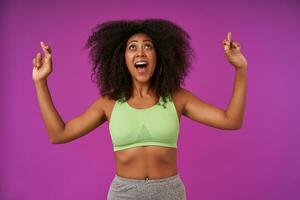 Cheerful young beautiful dark skinned woman with curls standing over purple background, crossing fingers for good luck and hoping for better, looking upwards with wide and happy smile photo