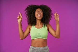 Indoor photo of attractive young dark skinned lady with curly hair wearing light green top, raising hands with crossed fingers for good luck, isolated over purple background with closed eyes