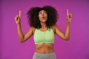 Indoor photo of young curly dark skinned female in light green top looking upwards scarely, posing over purple background with raised forefingers pointing above her head