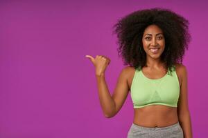 Positive young dark skinned woman with curly hair wearing sporty clothes, posing over purple background with raised hand and showing aside with thumb, looking at camera with happy and wide smile photo