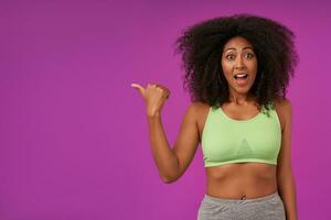 Indoor photo of overjoyed young curly woman with dark skin wearing casual hairstyle, wearing light green top over purple background, smiling widely to camera and raising eyebrows