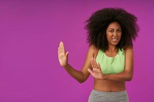 Indoor photo of young curly female with dark skin wearing casual hairstyle, frowning her face and rasing palms in refusing gesture, standing over purple background