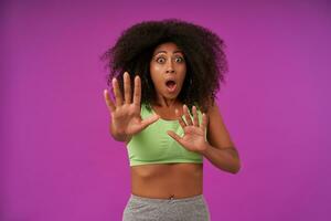 Portrait of frightened young dark skinned woman with curly hair wearing sporty clothes isolated over purple background, looking at camera with raised hands in stop gesture photo