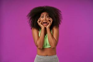 Pop-eyed young curly dark skinned woman standing over purple background with palms on her face, looking scarely to camera with wide mouth opened, wearing casual sporty clothes photo