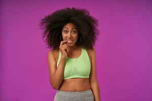 Pretty young dark skinned woman with curly hair wearing casual sporty clothes, looking to camera with raised eyebrows and keeping forefinger on underlip, standing over purple background photo