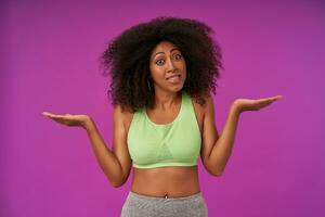 Puzzled young curly dark female with dark skin wearing light green top standing over purple background, looking at camera with confused face and shrugging photo