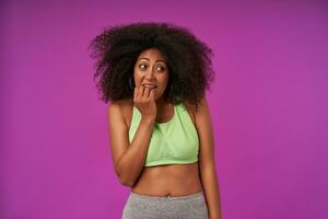 Indoor shot of attractive curly dark skinned lady with casual hairstyle wearing light green top, looking aside and covering mouth confusedly, isolated over purple background photo