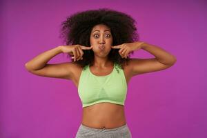 Funny indoor shot of young curly female woman with dark skin keeping forefingers on her face, while puffing out her cheeks, fooling over purple background in casual clothes photo