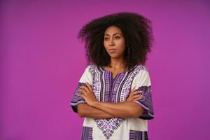 Studio photo on pretty young curly female with dark skin holding folded hands on her chest, looking aside with serious face, isolated over purple background in casual wear