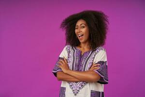 retrato de alegre Rizado oscuro desollado mujer con doblada manos sonriente extensamente y dando guiño a cámara, vistiendo blanco estampado camisa, posando terminado púrpura antecedentes foto