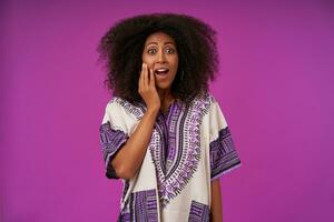 Inoor photo of amazed young dark skinned woman with curly hair keeping raised palm on her face and looking at camera with suprised face, isolated over purple background