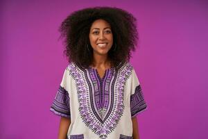 Attractive young curly lady with dark skin wearing white patterned shirt, posing over purple background with hands down, looking at camera with broad and sincere smile photo