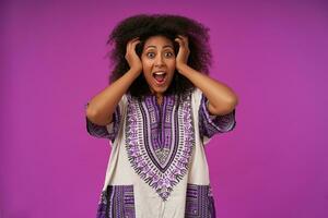 Overjoyed young dark skinned lady with curly hair wearing white patterned shirt, posing over purple with wide mouth opened and raising hands to her head photo