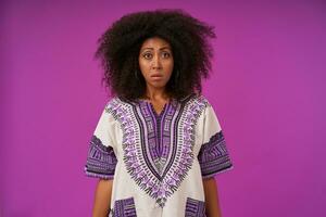 Portrait of upset curly dark skinned female in casual clothes standing over purple background wth hands down, contracting forehead and looking to camera with sad face photo