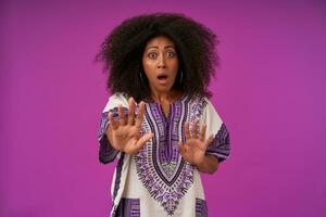 Horizontal shot of young curly dark skinned lady with casual hairstyle raising palms fearful while standing over purple background, looking at camera with raised eyebrows and round eyes photo