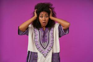 Scared young dark skinned woman with curly hair wearing white patterned shirt, holding her head with raised hands and looking at camera with wide eyes and mouth opened, isolated over purple background photo