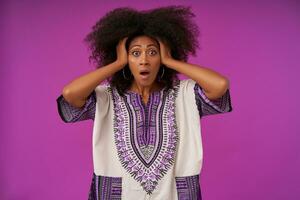 Studio shot of frightened young dark skinned female clutching her head with hands and looking to camera afraid, standing posing over purple background in casual clothes photo