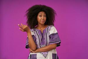 Portrait of young curly lady casual with dark skin in white patterned shirt rasing palm confusedly, looking at camera with raised eyebrows while standing over purple background photo