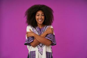 Happy young curly woman with dark skinn wearing white patterned shirt, posing over purple background with crossed hands on her chest, looking at camera joyfully with broad smile photo