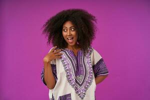 Joyful young dark skinned woman with casual hairstyle keeping palm on her shoulder and looking aside with wide joyful smile, posing over purple background photo