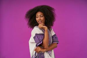 Young curly dark skinned woman wearing white patterned shirt touching face with raised hand, turning back and looking over shoulder with cheerful smile, posing over purple background photo