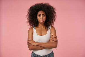 Portrait of offended young dark skinned woman with casual hairstyle standing over pink background with folded hands, looking at camera with pursed lips and upset face photo