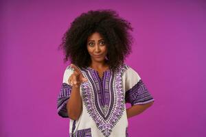 Portrait of unpleased young dark skinned female with hairstyle poiting aside with raised forefinger and folding lips, wearing white patterned shirt over purple background photo