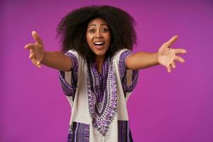 Overjoyed young pretty dark skinned female with casual hairstyle wearing white patterned shirt, posing over purple background with opened arms and going to give hug photo