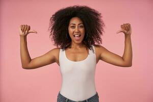 Happy young curly woman with dark skin wearing white shirt and jeans, raising hands and showing to herself with thumbs, posing over pink background with wide mouth opened photo