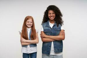 Studio shot of cheerful young dark skinned brunette female and redhead long haired pretty girl crossing hands while standing over white background and smiling happily to camera photo