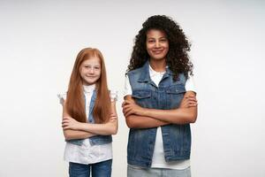 Two beautiful young long haired ladies folding hands on chest and looking positively at camera, being in nice mood and smiling sincerely, dressed in jeans vests and white shirts over white background photo
