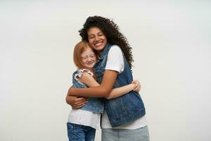 Embracing happy couple of young dark skinned curly brunette female and lovely red haired girl smiling broadly and keeping their eyes closed, isolated over white background photo