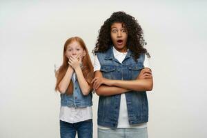 Open-eyed couple of young pretty ladies with long hair looking amazedly to camera and raising surprisedly eyebrows, dressed in casual clothes while posing against white background photo