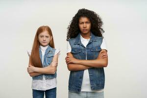 Severe young ladies with long hair standing over white background with folded hands, looking severely at camera and frowning eyebrows, dressed in casual family look photo