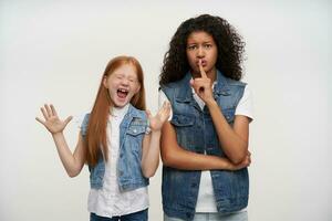 Horizontal shot of pretty young dark skinned curly brunette female holding index finger on her lips and asking to keep silence, standing over white background with excited foxy little girl photo