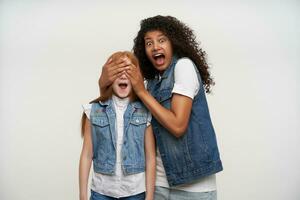 Por-eyed young dark skinned curly brunette lady screaming scaredly and covering eyes with palms to little long haired foxy girl, wearing casual clothes over white background photo