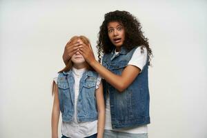 Indoor shot of scared dark skinned curly woman looking frightened at camera with wide mouth opened and closing eyes with raised hands to redhead little girl, isolated over white background photo