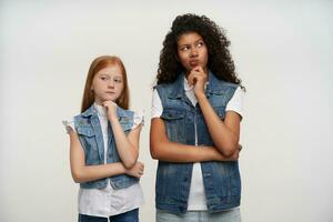 Two attractive pensive young ladies in jeans vests and white shirts looking thoughtfully in different sides and holding their chins with raised hands, standing against white background photo