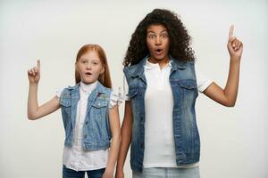 Open-eyed young couple of dark skinned curly female and foxy long haired little girl pointing up with forefingers and looking at camera with surprised faces, isolated over white background photo