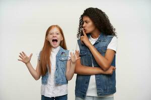 Studio photo of young curly brunette lady with dark skin raising hand in hush gesture and trying to calm down energized lovely redhead little girl, isolated over white background