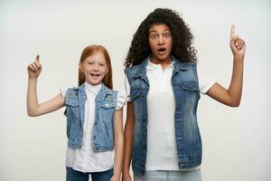 Surprised young lovely long haired girls in jeans vests and white shirts showing upwards with raised index fingers and looking at camera with wide mouth opened, isolated over white background photo